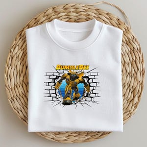 Könnte beinhalten: Weißes T-Shirt mit einem Aufdruck von Bumblebee, einem gelben Roboter aus der Transformers-Reihe, der eine Ziegelmauer durchbricht.