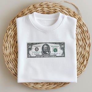 May include: White t-shirt with a printed image of a $50 bill featuring Abraham Lincoln. The bill has the serial number B 895060213 A.