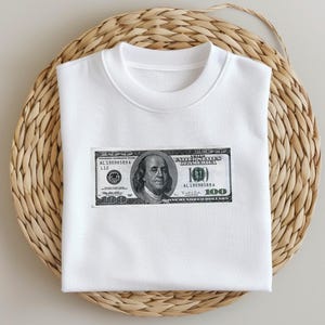 May include: White t-shirt with a graphic of a $100 bill featuring a portrait of Benjamin Franklin. The bill has the text "UNITED STATES OF AMERICA" and "ONE HUNDRED DOLLARS".
