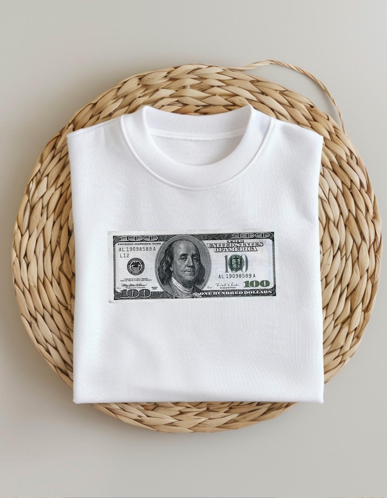 May include: White t-shirt with a graphic of a $100 bill featuring a portrait of Benjamin Franklin. The bill has the text "UNITED STATES OF AMERICA" and "ONE HUNDRED DOLLARS".