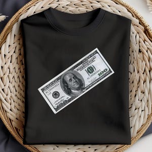 May include: Black t-shirt with a $100 bill printed on the front. The bill is positioned diagonally across the shirt. The bill is a US dollar bill with the portrait of Benjamin Franklin.