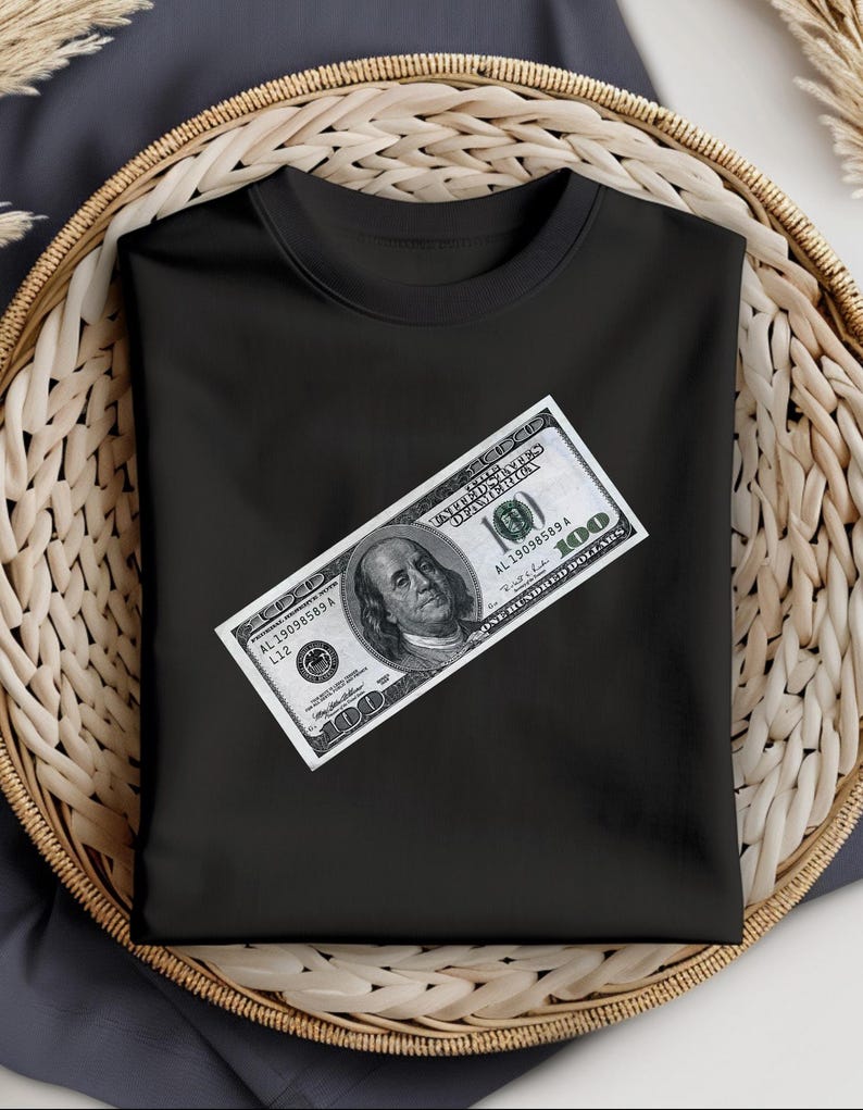 May include: Black t-shirt with a $100 bill printed on the front. The bill is positioned diagonally across the shirt. The bill is a US dollar bill with the portrait of Benjamin Franklin.