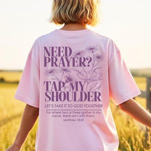 May include: A pale pink t-shirt featuring the text "NEED PRAYER? TAP MY SHOULDER" and a floral design. The shirt also includes the text "LET'S TAKE IT TO GOD TOGETHER" and a Bible verse. The shirt is worn by a person in a field.