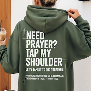 May include: Military green hoodie with the text "NEED PRAYER? TAP MY SHOULDER" in white. The hoodie also includes the text "LET'S TAKE IT TO GOD TOGETHER" and a Bible verse. The hoodie is a solid color and has a hood.