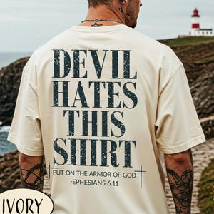 Puede incluir: Camiseta de color marfil con el texto "DEVIL HATES THIS SHIRT" en azul oscuro. Debajo, se lee "PUT ON THE ARMOR OF GOD - EPHESIANS 6:11". Una prenda de vestir informal.