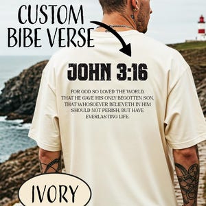 May include: Ivory t-shirt featuring the text "JOHN 3:16" and a Bible verse on the back. The shirt also has the words "CUSTOM BIBE VERSE" above the text. The shirt is being worn outdoors.