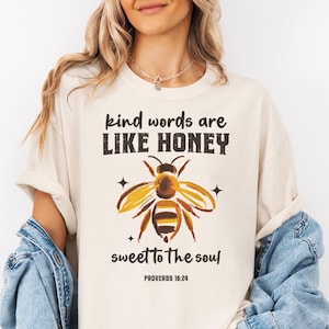 Może przedstawiać: Kremowa koszulka z napisem "kind words are like honey sweet to the soul" i grafiką pszczoły. Pszczoła jest żółto-brązowa z czarnymi paskami. Tekst "Proverbs 16:24" znajduje się poniżej.