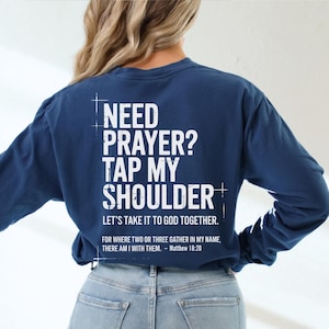 May include: Navy blue long-sleeve shirt with white text on the back that reads "NEED PRAYER? TAP MY SHOULDER. LET'S TAKE IT TO GOD TOGETHER." Below the text is a Bible verse. The shirt is a comfortable fit.