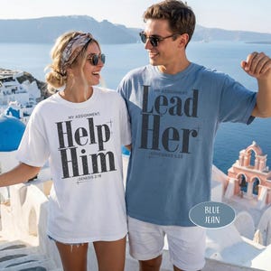 May include: Two people wearing matching t-shirts with text. The white shirt says "Help Him" and the blue shirt says "Lead Her". Both shirts have additional text below. The background shows a coastal scene with white buildings and blue domes.