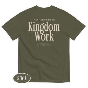 May include: Sage green t-shirt with the text "FATHERHOOD IS Kingdom Work" in a cream-colored font. The text "PROVERBS 22:6" is below the main text. A small oval label at the bottom left says "SAGE".