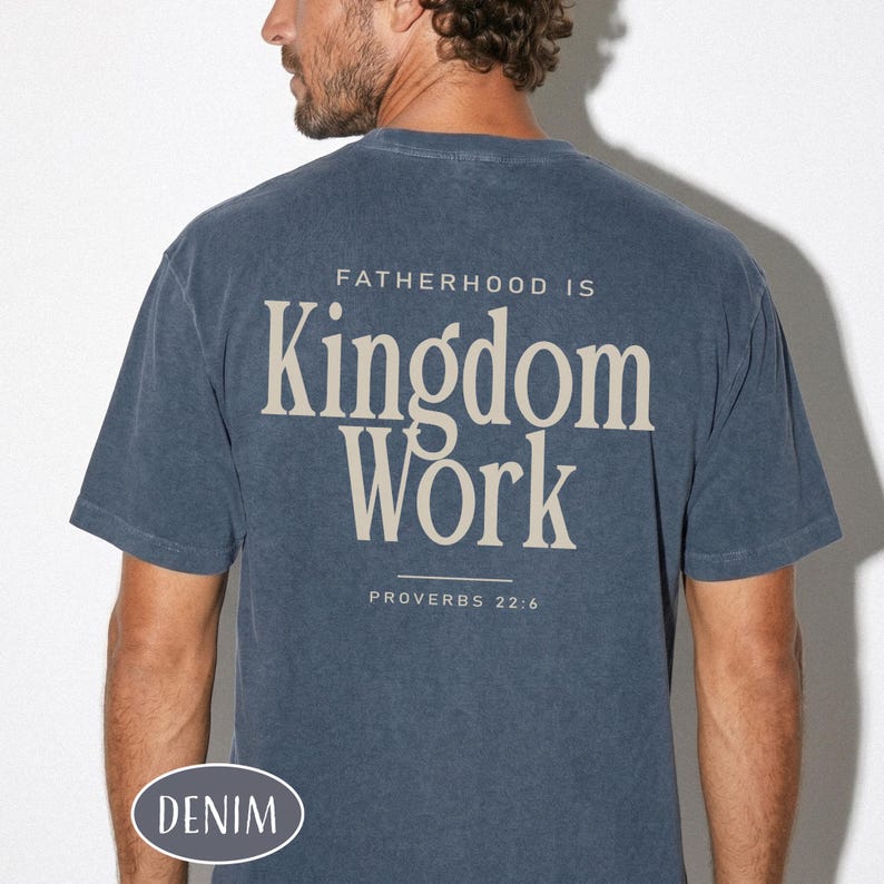 May include: A denim blue t-shirt with the text "FATHERHOOD IS Kingdom Work" in a cream color. The text "PROVERBS 22:6" is below the main text. The word "DENIM" is printed on the bottom left of the shirt.