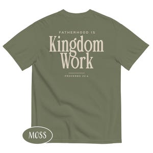 May include: A moss green t-shirt with the text "FATHERHOOD IS Kingdom Work" in a cream-colored, bold font. The text "PROVERBS 22:6" is below the main text. The shirt has a crew neck and short sleeves.