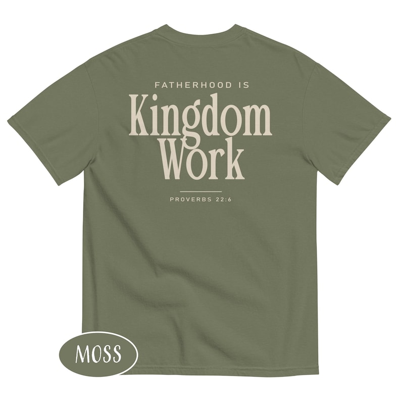 May include: A moss green t-shirt with the text "FATHERHOOD IS Kingdom Work" in a cream-colored, bold font. The text "PROVERBS 22:6" is below the main text. The shirt has a crew neck and short sleeves.