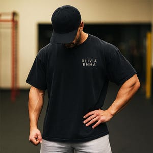 May include: Black t-shirt and baseball cap. The t-shirt has the text "OLIVIA EMMA" printed on the front. The person is wearing a black baseball cap. The background is a gym setting.