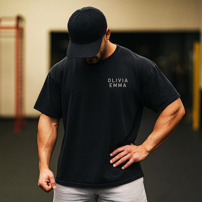 May include: Black t-shirt and baseball cap. The t-shirt has the text "OLIVIA EMMA" printed on the front. The person is wearing a black baseball cap. The background is a gym setting.