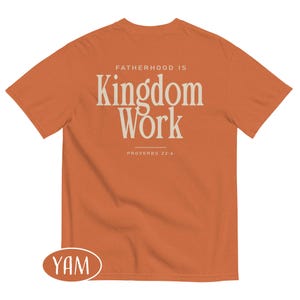 May include: An orange t-shirt with the text "FATHERHOOD IS Kingdom Work" in a cream-colored font. The text "PROVERBS 22:6" is below the main text. The shirt is a short-sleeved crew neck.