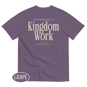 May include: A grape-colored t-shirt with the text "FATHERHOOD IS Kingdom Work" in a cream-colored font. The text "PROVERBS 22:6" is below the main text. The word "GRAPE" is in a white oval at the bottom left.