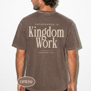 May include: Espresso-colored t-shirt with the text "FATHERHOOD IS Kingdom Work" and "PROVERBS 22:6" printed on the back. The shirt is a solid color and has a crew neck. The word "ESPRESSO" is printed on the bottom left.