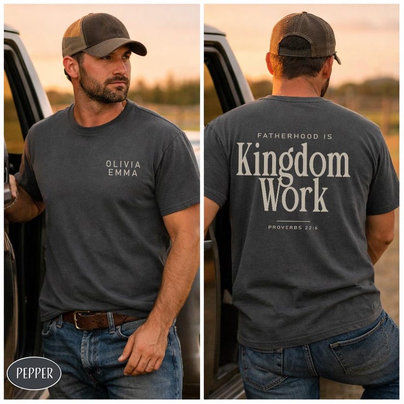 May include: Dark gray t-shirt with the text "OLIVIA EMMA" on the front and "FATHERHOOD IS Kingdom Work PROVERBS 22:6" on the back. The person is wearing a brown and tan baseball cap and blue jeans.