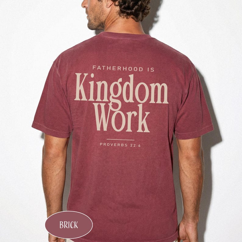 May include: Brick-colored t-shirt with the text "FATHERHOOD IS Kingdom Work" and "PROVERBS 22:6" printed on the back. The shirt is a solid color and has a crew neck. The word "BRICK" is printed on the bottom of the shirt.