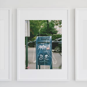 May include: Framed art print featuring a blue mailbox with graffiti and a whimsical outline of a figure. The background shows a street scene with cars and trees, all within a white frame.