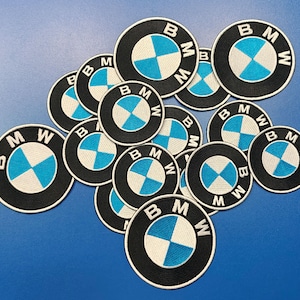 May include: A collection of round embroidered patches featuring the BMW logo. Each patch has a black border with the letters "BMW" in white, surrounding a blue and white segmented circle. The patches are scattered on a blue surface.
