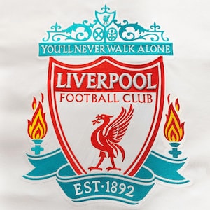 Könnte beinhalten: Ein rotes und weißes Wappen mit einem Vogel und den Worten "Liverpool Football Club" und "You'll Never Walk Alone". Das Wappen ist von einem blauen und weißen Band mit den Worten "EST. 1892" umgeben.