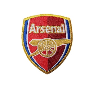 Op de afbeelding: Geborduurde patch met het wapen van voetbalclub Arsenal. Het wapen is schildvormig, met een rode en blauwe achtergrond, gouden rand en het woord "Arsenal" in het wit. Een gouden kanon is gecentreerd op het wapen.