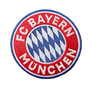 Może przedstawiać: Haftowana naszywka z logo FC Bayern Monachium. Okrągła naszywka ma czerwoną obwódkę z białym napisem "FC BAYERN MUNCHEN". Środek przedstawia biało-niebieski wzór w romby.