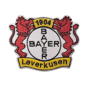 Może przedstawiać: Haftowana naszywka z logo Bayer Leverkusen. Naszywka ma białe tło z okrągłym emblematem pośrodku, słowem "BAYER" w kolorze czarnym i dwoma czerwonymi lwami po obu stronach. Obecne są również teksty "1904" i "Leverkusen".