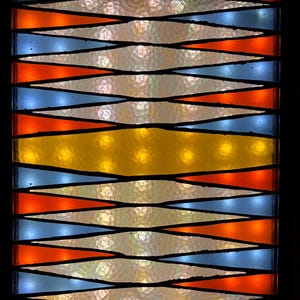 May include: A stained glass panel with a geometric design. Alternating rows of orange and blue triangles are separated by black lines. A yellow horizontal band is in the center. The background is a textured, off-white color.