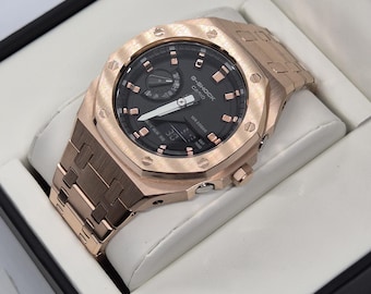 May include: A rose gold G-Shock Casio watch with a black face and digital display. The watch is in a black box with a white interior. The watch has a metal band and a round face with a textured bezel.