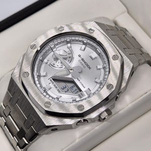 May include: A silver G-Shock Casio watch with a stainless steel strap. The watch face is silver with black detailing, showing the time and date. The watch has a square bezel with visible screws, presented in a white box.
