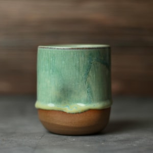 Könnte beinhalten: Ein handgefertigter Keramikbecher mit einer einzigartigen Glasur. Der obere Teil ist ein leuchtendes Türkis, das in ein hellgelbes Band übergeht und mit einem warmbraunen Sockel endet. Der Becher ist zylindrisch und hat eine glatte Oberfläche.