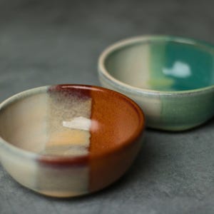 May include: Two small, handmade ceramic bowls. One bowl is divided into cream, brown, and orange sections. The other bowl is divided into cream and teal sections. Both bowls have a glossy finish and are set against a gray background.