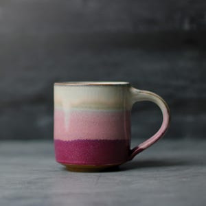 May include: A ceramic mug featuring a horizontal gradient design that transitions from a creamy off-white at the top to a soft pink in the middle and a deep magenta at the base. The mug has a curved handle and a smooth, speckled texture throughout.