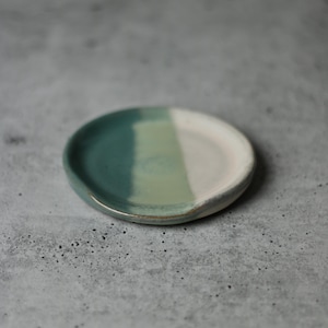 Può includere: Un piccolo poggia-cucchiaio rotondo in ceramica. Il lato sinistro è verde acqua, che sfuma in verde chiaro e poi in bianco a destra. Il poggia-cucchiaio ha una leggera rientranza per tenere le posate. Diametro di circa 10 cm.