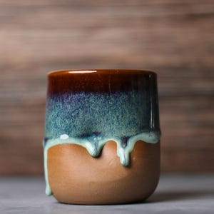 May include: A handcrafted ceramic tumbler with a distinctive glaze. The top is a deep brown, fading to teal and dripping down the sides over a tan base. The cup has a smooth, rounded form.