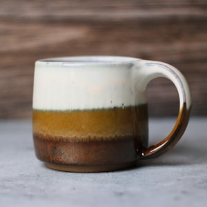 May include: A ceramic mug with a layered color scheme. The top section is white, followed by a band of olive green, then a band of tan, and the bottom is brown. The handle is brown. The mug is on a gray surface.