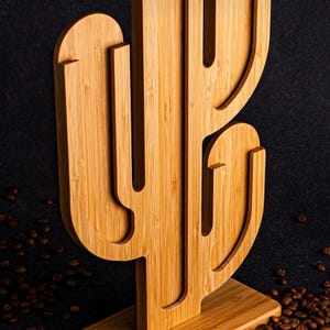 Può includere: Un portacapsule per caffè in legno a forma di cactus con una base. Il portacapsule è realizzato in bambù marrone chiaro e ha una finitura in legno naturale. Il portacapsule è posto su una superficie ricoperta di chicchi di caffè.