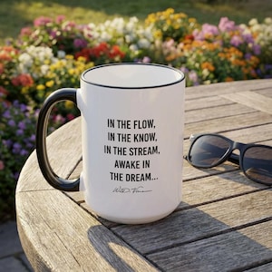 Puede incluir: Taza de cerámica blanca con asa y borde negros. La taza presenta el texto "IN THE FLOW, IN THE KNOW, IN THE STREAM, AWAKE IN THE DREAM..." y una firma. Gafas de sol negras sobre una mesa de madera.