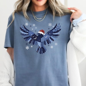 May include: Blue jean colored t-shirt featuring a stylized bird design with a Santa hat. The bird is composed of various blue patterns and surrounded by snowflakes. The text "BLUE JEAN" is at the bottom of the image.