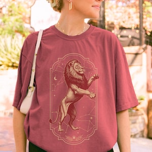 May include: A crimson short-sleeved t-shirt featuring a central graphic of a standing lion within an ornate rectangular frame. The design includes thin white lines depicting constellations and a crescent moon, set against the solid red fabric.