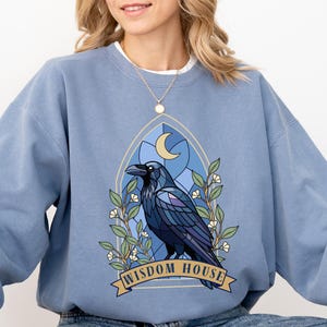 May include: A light blue sweatshirt featuring a raven design within a stained-glass window, with the text "WISDOM HOUSE" on a banner. The raven is detailed with shades of blue and purple, surrounded by floral accents and a crescent moon.