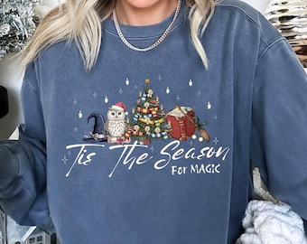 May include: A blue sweatshirt with the text "Tis The Season For Magic". The graphic features a Christmas tree, an owl wearing a Santa hat, a witch hat, a book, and a broom. The sweatshirt is a comfortable, casual style.