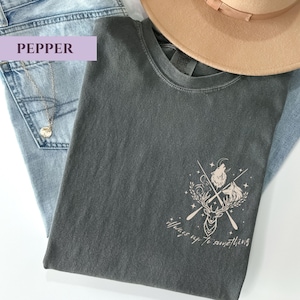 May include: A dark grey t-shirt featuring a deer, wolf, and crossed oars graphic, with the text "Always up to something." The shirt is on light blue jeans, a gold necklace, and a beige hat.