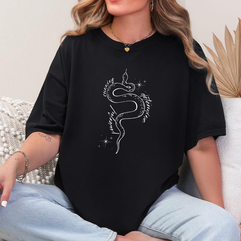 May include: A black t-shirt with a white snake design. The snake is curled up and has the words 'cunning', 'powerful', 'determined' written around it.