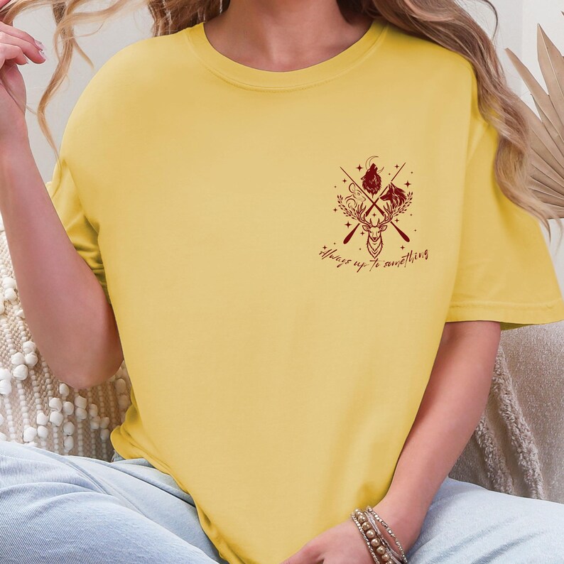 May include: Mustard yellow t-shirt with a maroon graphic. The graphic includes a deer head, crossed arrows, and the text "Always up to something". The shirt has a crew neck and short sleeves.