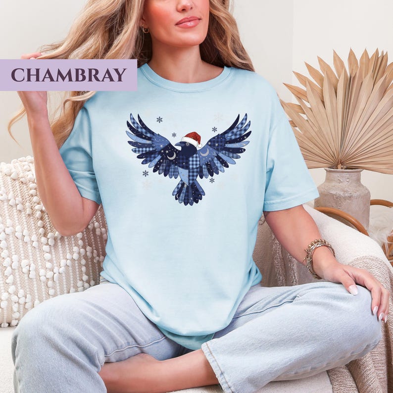 May include: Light blue t-shirt with a graphic of a bird wearing a Santa hat. The bird design features various blue patterns and snowflake accents. The word "CHAMBRAY" is printed in a lavender rectangle in the upper left corner.