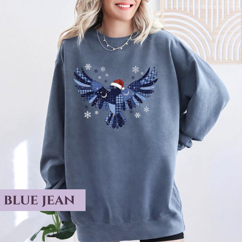 May include: Blue jean colored sweatshirt featuring a graphic of a bird with outstretched wings, adorned with snowflakes and a Santa hat. The bird design incorporates various blue patterns. The text "BLUE JEAN" is displayed at the bottom.